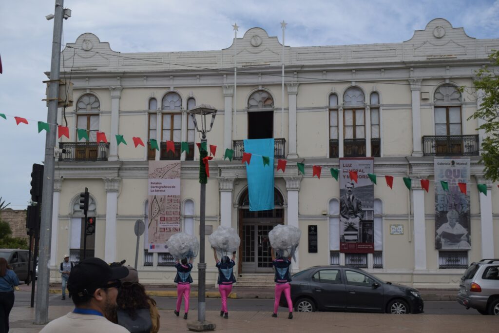Fotografía de la obra "Siluetas de Agua", de la Compañía Teatro en Movimiento Callejerx. En ella se puede ver a 3 intérpretes esperando que caiga una tela celeste desde una de las ventanas del Museo Gabriel González Videla, en La Serena