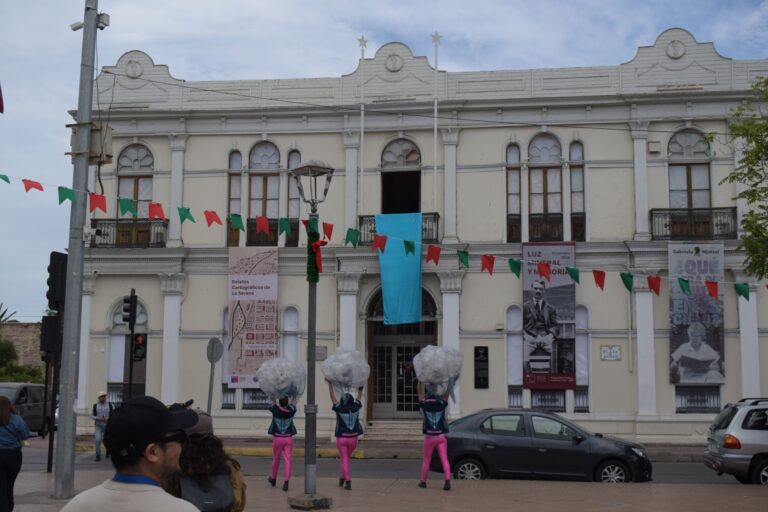 Fotografía de la obra "Siluetas de Agua", de la Compañía Teatro en Movimiento Callejerx. En ella se puede ver a 3 intérpretes esperando que caiga una tela celeste desde una de las ventanas del Museo Gabriel González Videla, en La Serena